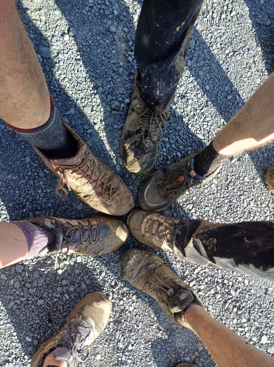 Group of hikers standing in a circle, each extending one muddy hiking boot toward the centre after completing the Dome Valley to Govan Wilson track in New Zealand