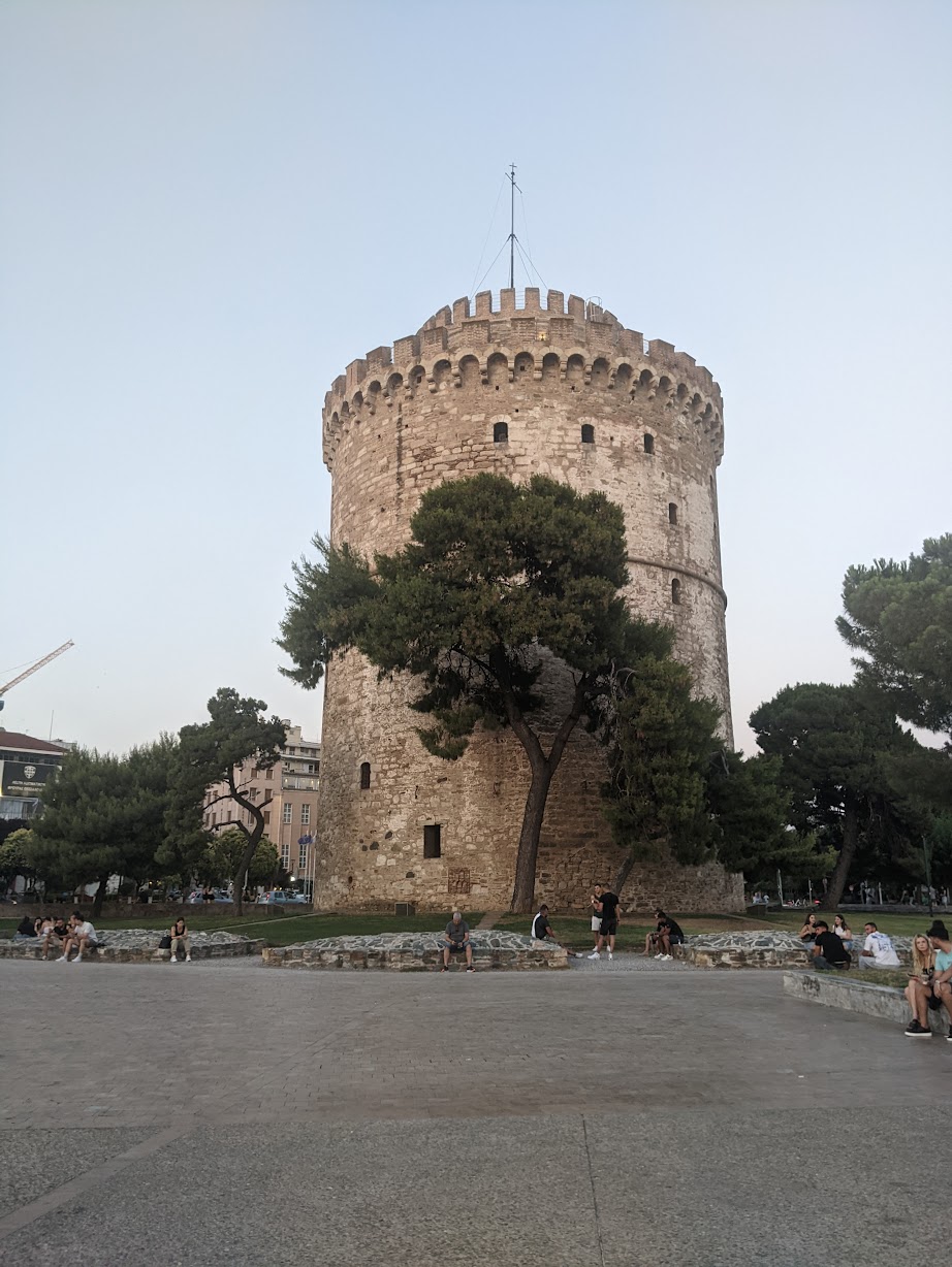 The White Tower of Thessaloniki at dusk, with people sitting on the grass around its base