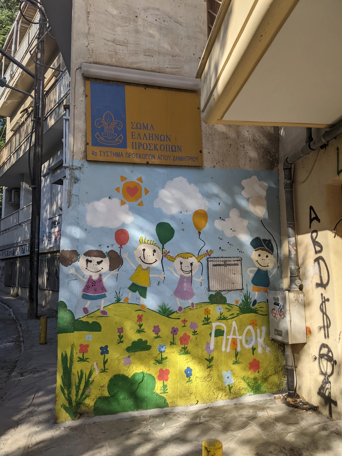 Children's mural with figures, balloons and flowers beside a Greek scout-troop sign, Thessaloniki