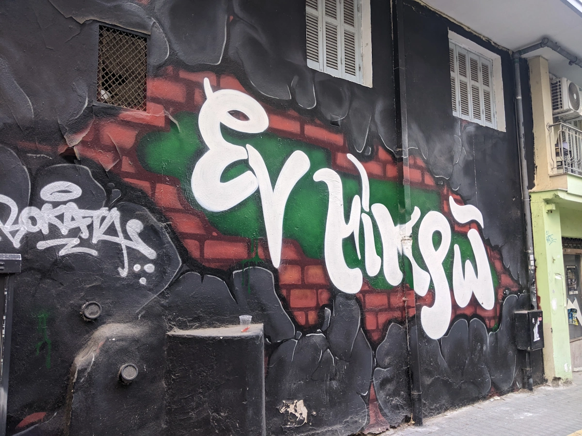 Black wall graffiti with white script lettering over a painted brick design, Thessaloniki